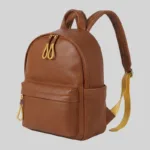 Three-quarter angle of premium top-grain leather wholesale daypack highlighting side slip pockets, gold-tone hardware, and contrast yellow webbing.