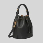 Side profile of structured black top-grain cowhide bucket bag featuring top handle rivets and detachable crossbody strap.